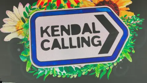 A sign for Kendal Calling. It looks like a road sign pointing right, and there are flowers and vines entwined around it.
