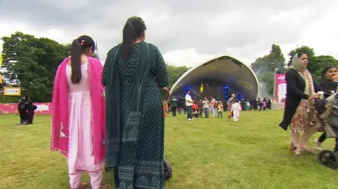Two people are prominent in the photo, standing up on grass with their backs to the camera. More than a dozen people are in the background.