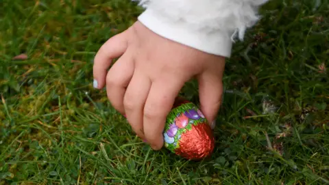 Getty Images A child's hand is picking up a foil-wrapped chocolate egg from a patch of grass. 