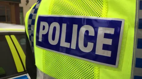 The back of a police officer wearing a bright yellow-high vis jacket stating 'police' in capitals in silver and blue. The side of a police car is on the left.