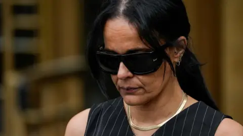 A woman wearing dark sunglasses walks with her head pointed down.