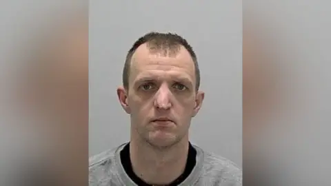 Bedfordshire Police The head and shoulders of a man with short dark hair wearing a black, round-necked T-shirt under a grey round-necked top.