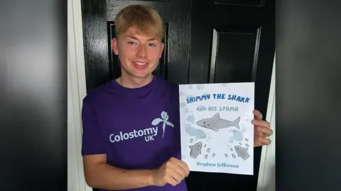 Stephen Jefferson holding a copy of his book Shimmy the shark and his stoma. The book cover is white, with a blue title and several sharks. Mr Jefferson is smiling at the camera and wearing a purple Colostomy UK t-shirt. He has blond hair and blue eyes.