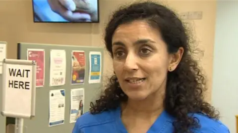 A woman with dark curly hair in blue scrubs stands in front of a notce board looking in to the camera.