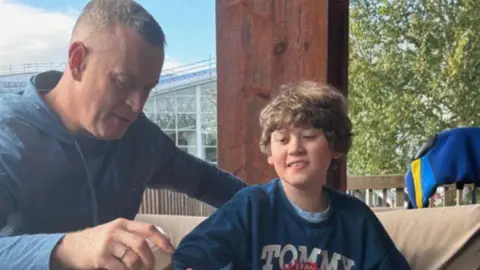 Deirdre Shakespeare Harry is sitting with his dad. Harry has brown curly hair and is wearing a navy sweatshirt that says the word Tommy in white writing. His dad has a navy hoodie on and has short, brown hair.