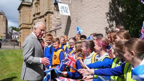 Getty Images The King meeting local children. They are waving flags and looking happy and he is chatting away to them. Jedburgh Abbey can be seen in the background.