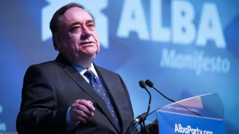 PA Media A dark haired main, Alex Salmond, wearing a dark suit and blue tie, stands at a podium with Alba in the background 