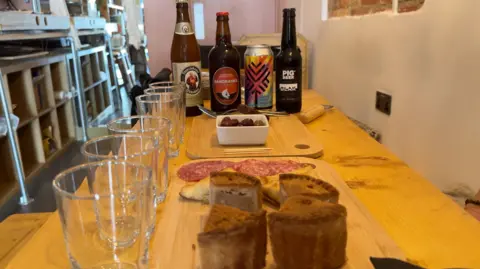 Image shows beers and food on a table ready to be paired. In the background, there are four local beers. Two bottles then a can and then a fourth bottle. There are six glasses lined up along the table too. There are two wooden boards on the table with a pot of olives, salami, samosas and pork pies.