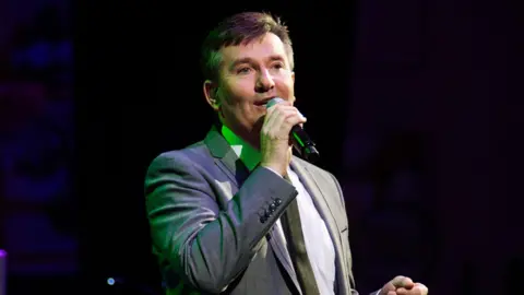 Daniel O'Donnell is wearing a grey suit jacket, a white shirt and a black tie as he sings into a microphone. He is performing on stage, with a green light just behind him reflecting off his brown, short hair.