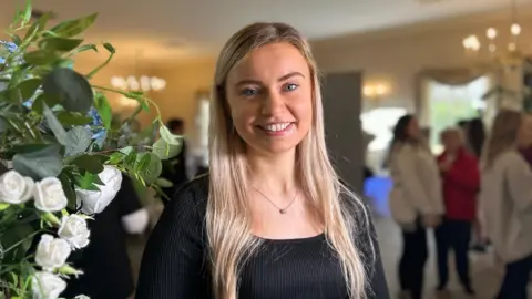 Meg Rolley is a young woman with long blonde hair and blue eyes. She is smiling and looking straight ahead. She is wearing a black top and a necklace with a fine chain and pendant.