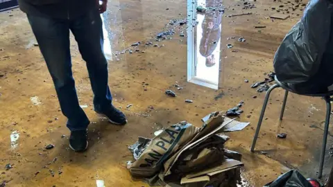 There is water, dirt and soggy cardboard on the floor of a dance studio. In the image you can also see a man's shoes.