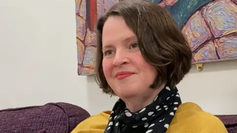 A white woman with brown hair and brown eyes, wears a black spotty scarf, yellow jumper and black leggings. She sits on a purple sofa in front of a colourful art canvas.
