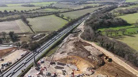 Shaun Whitmore/BBC An aerial image of a dual carriageway with cars travelling along it, with fields in the near and far distance. There is an extensive excavation area in the right-hand corner of the photo.