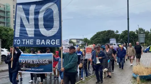 BBC News Dozens of people marching down the street, holding a large banner which reads "no to the barge"