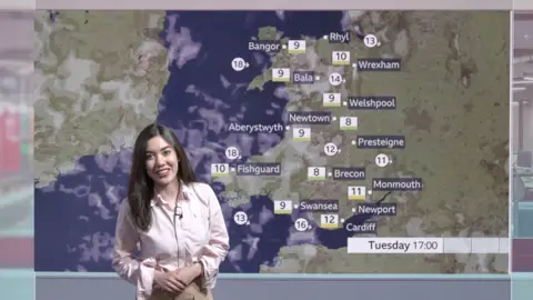 BBC Wales' Sabrina Lee standing in front of a weather map showing the whole of Wales and the various temperatures. Sabrina has long dark hair and is wearing a pink shirt with rolled up cuffs and a clip microphone is visible on her shirt