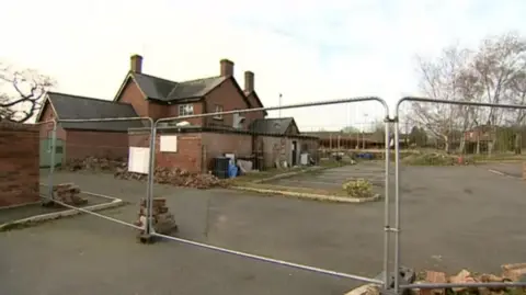 A large brick building with a car park in front of it and metal fencing around the outside