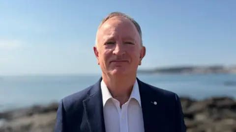 Man stood in front of coastline wearing a blue blazer and a white shirt.