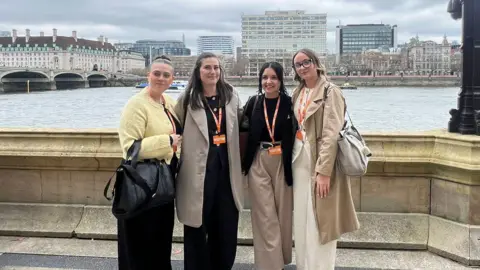 Faye Ramsey Four women stand on the bank of the River Thames at Westminster. Behind them parts of London can be seen on a grey cloudy day.