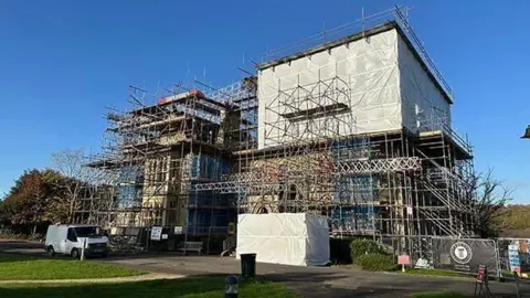 Mole Valley District Council A large building is covered by scaffolding. Some of the scaffolding is wrapped in protective sheeting. A white van is parked in front,