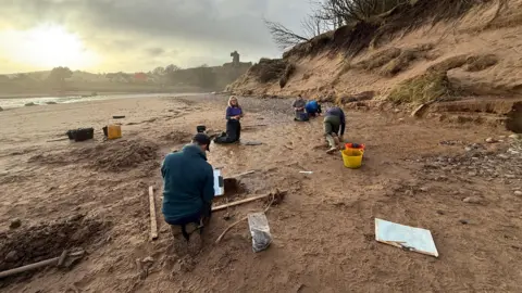 University of Aberdeen A wide scene showing University of Aberdeen archaeologists working on the discovery of ancient footprints on the beach at Lunan Bay, Angus, with the North Sea to the left and dunes to the right, in weak winter sunshine. 