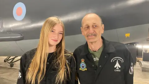 Grace, who has straight, long, blonde hair, wearing black overalls and smiling at the camera. She is stood next to her grandad Chris who has a grey moustache and is wearing a green t-shirt and black overalls. They are stood in front of an aircraft.
