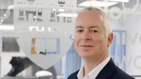 Dr Michael Sykes. He has short grey hair and is wearing a suit. He is standing in front of a glass window embossed with the university's emblem.