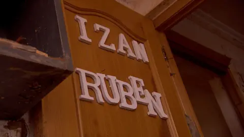 A broken wooden door with white letters spelling out words "Izan" again "Reuben" nailed to it