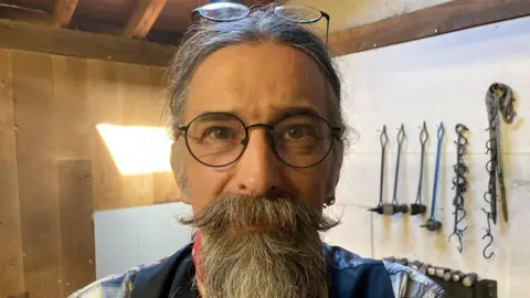 BBC/Tom Percival John Wills with short hair and a long beard standing in a workshop