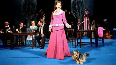 A small Yorkshire terrier-type dog on stage during a performance of La Boheme. The dog's lead is being held by the character of Musetta who is wearing a long cerise dress with a wide white fur collar. She has long auburn hair. Behind her are some people sitting at cafe tables.