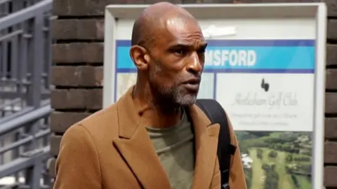 Matthew Martin in a light brown coat and green T-shirt, carrying a black rucksack 