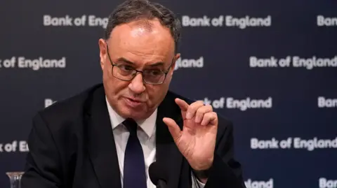 Bank of England governor Andrew Bailey sitting in front of a Bank of England backboard and gesturing with his hand