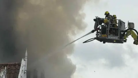 Using an aerial ladder, fire crews tackle a fire. They are using a hose and there is a large plume of smoke. 