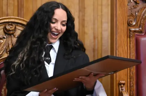 Jade Thirwall looking down at the picture she has been presented by South Tyneside Council. She is has long black hair and is wearing a black blazer and tie with a white shirt. She has her mouth wide open in a smile.