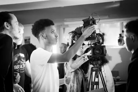Compass Collective This black and white image shows a group of young people preparing equipment ready for filming. There is a camera on a tripod with people gathered round making adjustments.