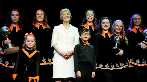 Jordan Pettitt/PA Wire Catherine Connolly is stood on a stage wearing a white suit jacket and white skirt. She is smiling and is surrounded by Irish Dancers who are wearing black Irish dancing dresses with orange collars. 
