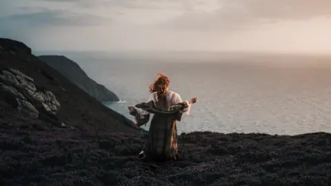 Ciara stands wearing traditional Celtic clothing on the edge of a cliff looking out to sea.
