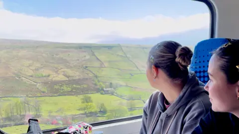 Nick Wooley/BBC Two young women look out of the window to see green rolling hills