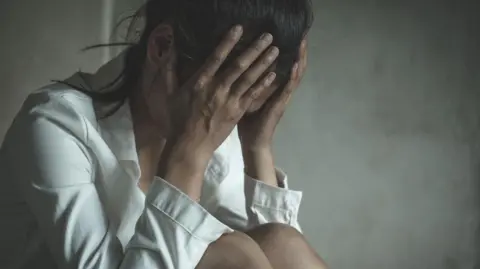 A woman with her head in her hands. She has dark hair which is tied back and she is wearing a white shirt.