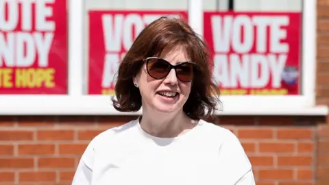 Reuters Lucy Powell, a woman in white T-shirt, with shoulder length hair and sunglasses, pictured against a backdrop of Vote Andy posters