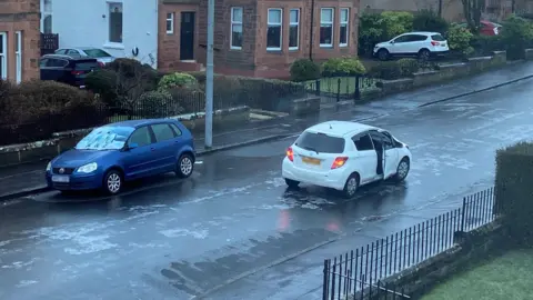 White car stuck in road unable to move forward or back due to ice which is visible on the road. The car door is open. There is a parked blue car near it and houses at the side of the road. 