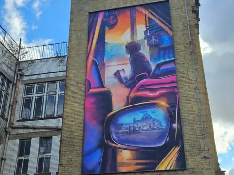 Aisha Iqbal/BBC The mural depicts a young woman reading while she leans against a car with a cityscape reflected in her car’s wing mirror.