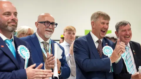 PA Media Reform councillors (left to right) Bradley Thompson, Mark Webster, Peter Harris and Jeff Bray. The first three are all in navy suits. They are clapping, standing in a hall, wearing blue rosettes.