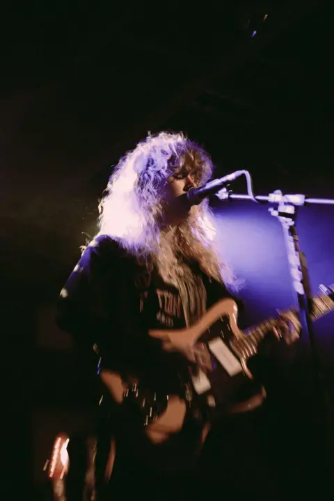 Jess Greaves A black background with a young woman with blonde curly hair and a full fringe holding an electric guitar and singing into a mic. There is a purple light shining on her.