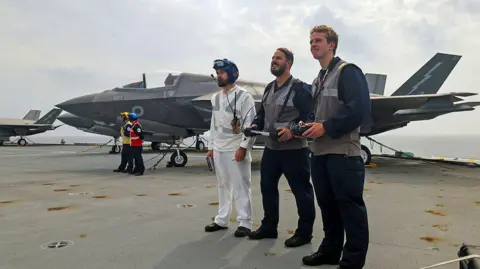 PO Phot Chris Sellars Three men stand to the right, two have large consoles in their hands, attached to lanyards around their necks and next to them is a man dresssed in white with a helmet. Behind them are jets on the deck of a frigate. 