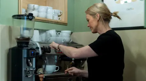 Sara seen from the side wearing a black t-shirt and blonde hair in a pony tail operating a large espresso machine in a cafe. Cups are stacked on top of the machine.