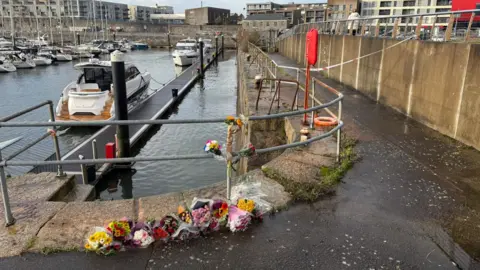 Floral tributes around Millbay Marina in Plymouth. There is water in the marina with boats stationary on the water. There is a police cordon in the background.