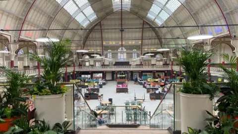 A view inside Derby Market Hall, looking down from the first floor to the ground floor. Plants are placed either side of a staircase with stalls visible in the background.