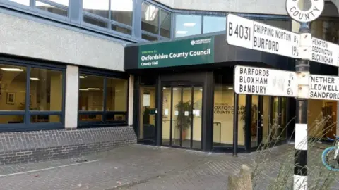 A view of the front entrance to Oxfordshire County Council's office building, County Hall. Its green sign is seen above some automatic doors, and road signs to nearby towns are pictured nearby.