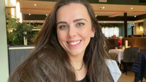 BBC Hanan Baba is a young woman with brown hair. She is smiling, and can be seen sitting in a restaurant and looking directly at the camera, with a visible earring and necklace.