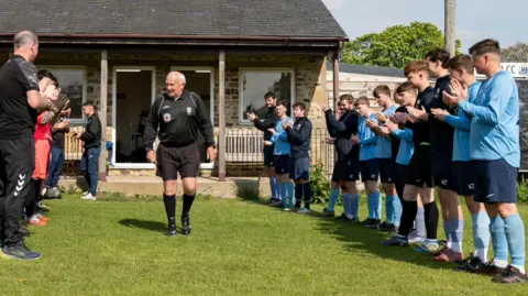 LS Photography The image shows an outdoor scene on a sports field. In the background there is a small stone clubhouse with a dark roof, a doorway, and windows. In the center a man dressed in a black referee-style uniform is walking forward across the grass, between two lines of players facing inward on either side, creating a guard of honour. On the right side, a group of young men wear light blue football kit. On the left side is a smaller group of individuals in darker sportswear, including black and some red clothing. Both groups are clapping, with hands raised in front of their bodies.